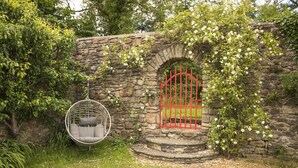 A stone wall with a red gate, a hanging chair, and white flowers. - 18th Century Mill House in a Courtyard Setting (.)