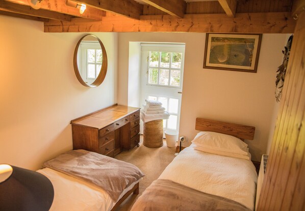 A bedroom with two beds, a wooden dresser, a round mirror, and a window with a view of greenery.