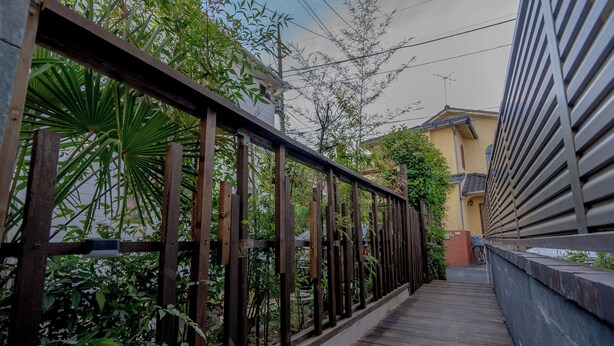 Un sendero de madera con una cerca de metal, rodeado de follaje y un edificio con techo inclinado.
