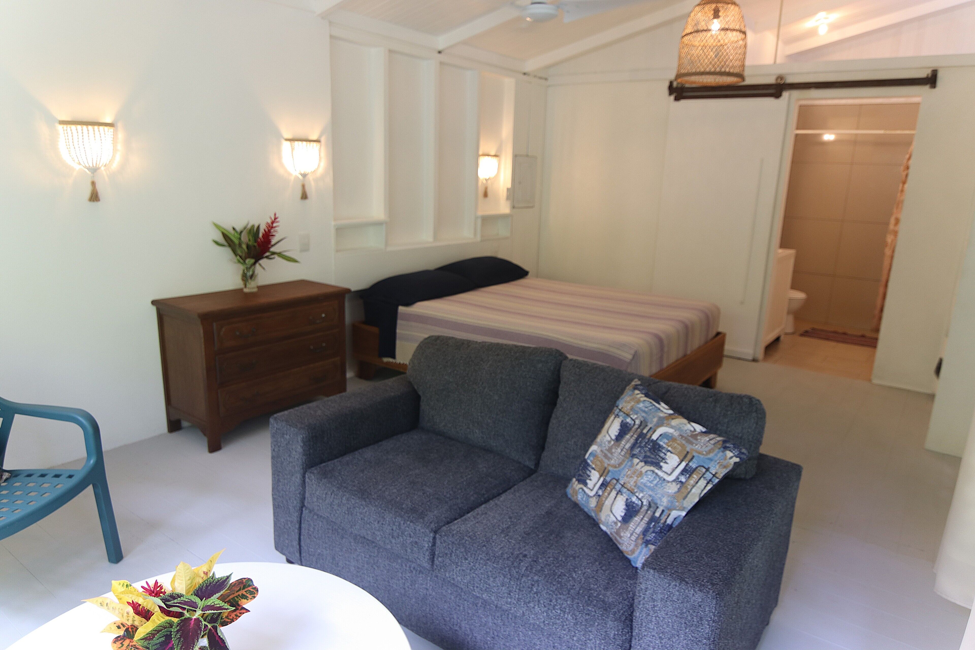 A bedroom with a bed, a grey sofa, a wooden dresser, and a small table with a plant.