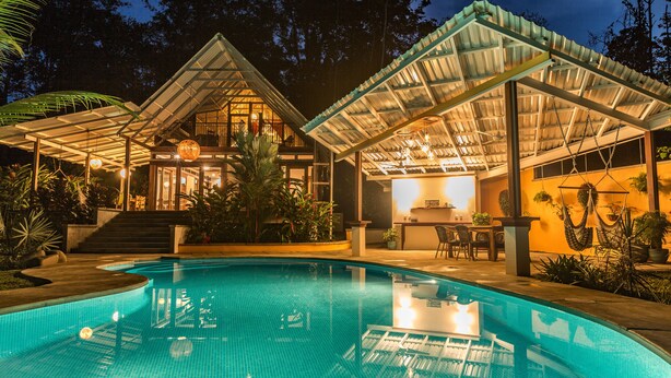 Una piscina con un porche cubierto, rodeada de abundante vegetación y un espacio interior bien iluminado.