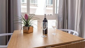 Een houten tafel met een fles wijn en twee glazen, een bloempot en uitzicht op een balkon door een raam met gordijnen.