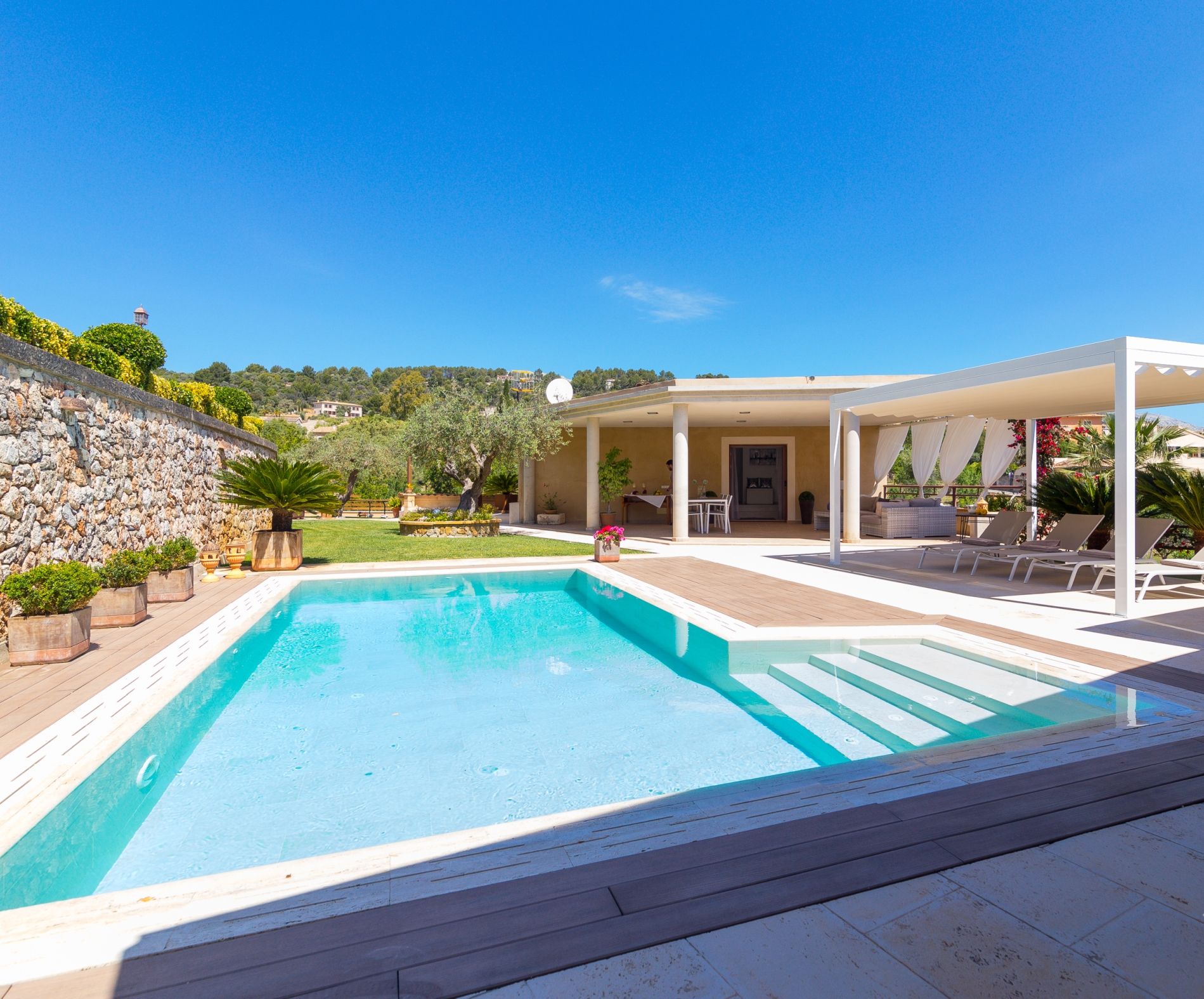 Une piscine rectangulaire entourée d’une terrasse en bois, un espace extérieur aménagé avec des meubles blancs et abrité, ainsi qu’un mur de pierre sur l’un des côtés.