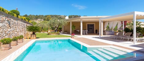 A rectangular swimming pool with a wooden deck surrounding it, a covered outdoor seating area with white furniture, and a stone wall on one side.