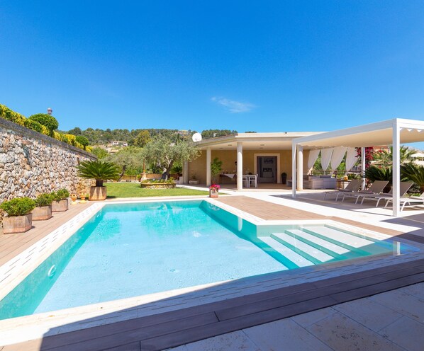 A rectangular swimming pool with a wooden deck surrounding it, a covered outdoor seating area with white furniture, and a stone wall on one side.