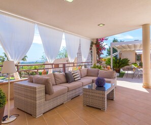 A spacious outdoor seating area with a large beige sofa and a glass coffee table, surrounded by white curtains and potted plants.