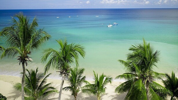 Una playa con agua turquesa cristalina, palmeras y barcos.