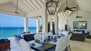 Un espacio cubierto para comer al aire libre con vista al océano, muebles blancos y ventiladores de techo.