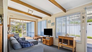Una sala de estar acogedora con sofá, televisión, escritorio de madera y ventanas amplias con persianas.