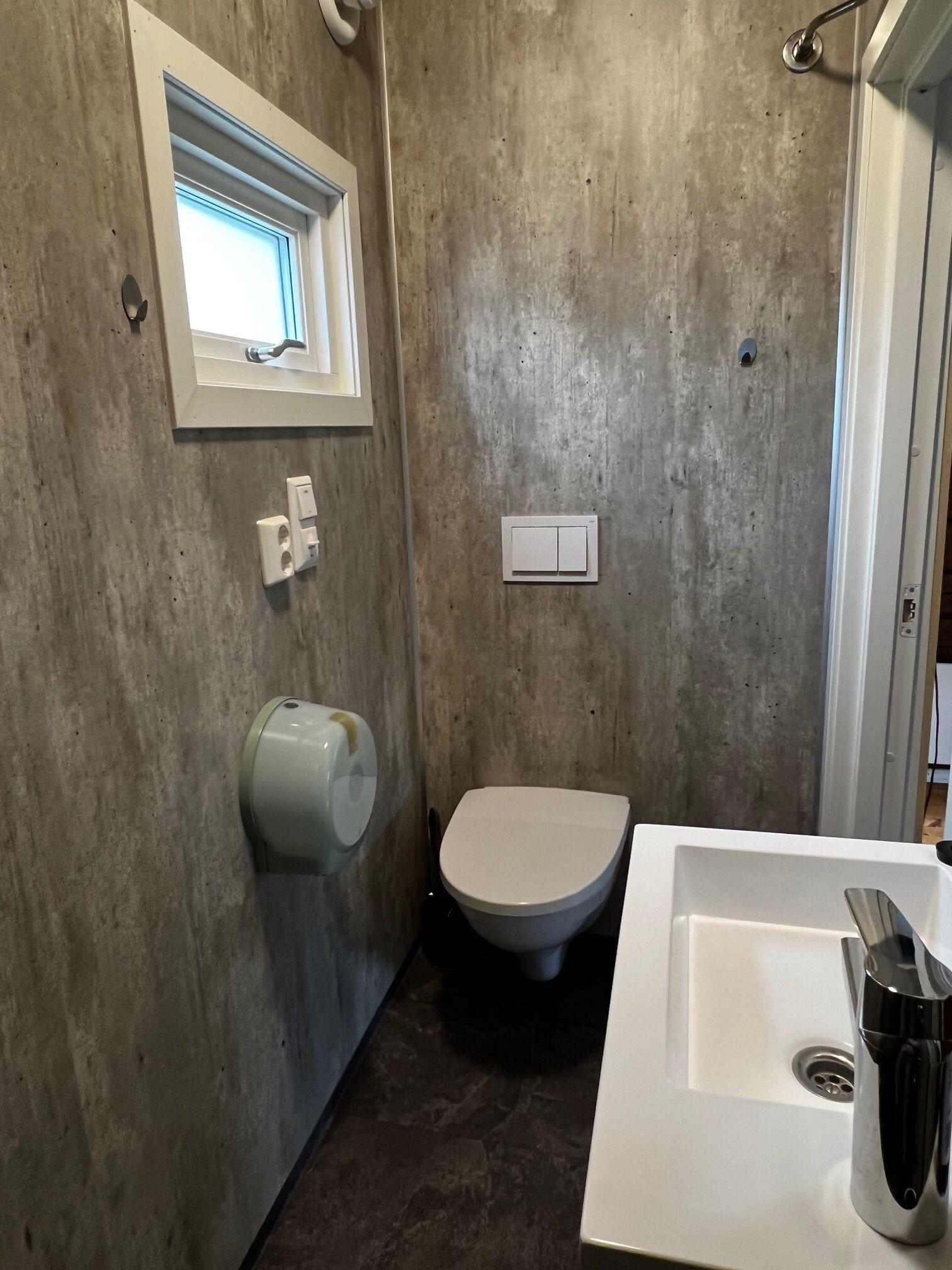 A bathroom with a concrete wall, a window, a toilet, a sink, and a hand dryer.