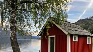 A small red wooden cabin with a green door, located by a lake with mountains in the background.