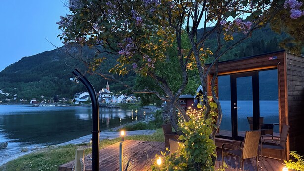 A wooden cabin with a deck overlooking a lake, surrounded by greenery and a distant village.