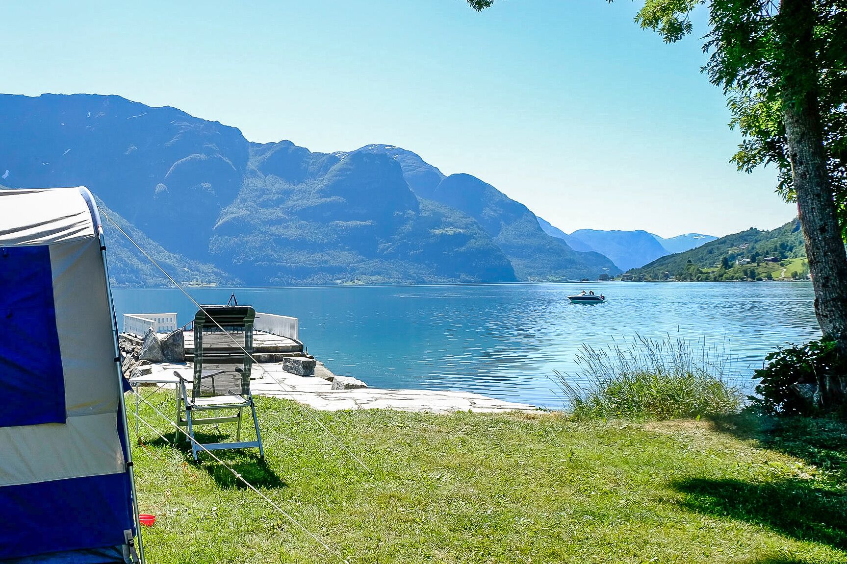Ein Campingplatz am Seeufer mit Zelt, Liegestuhl und Boot auf dem Wasser.