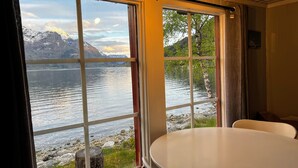 A room with a large window offering a view of a lake and mountains. There is a round table and a chair in the room.