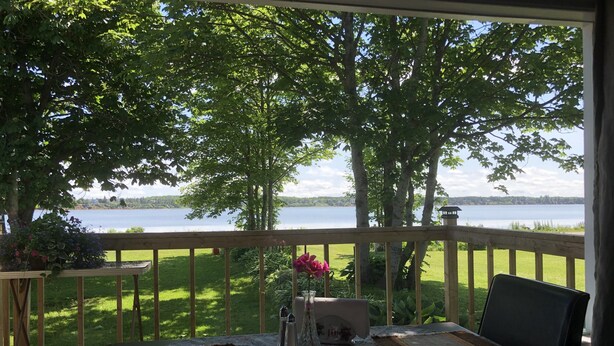 A room with a view of a lake, a wooden deck, and a table with a rug and a vase.