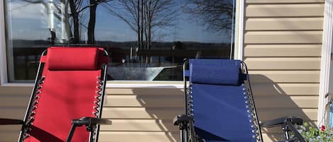 Dos sillones de exterior con estructura metálica y cojines rojo y azul, ubicados en una terraza de madera frente a una casa con revestimiento color beige y un amplio ventanal que refleja árboles y un cielo despejado.