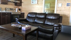 A cozy living room with a leather sofa, a coffee table, and a kitchen area with wooden walls and cabinets.