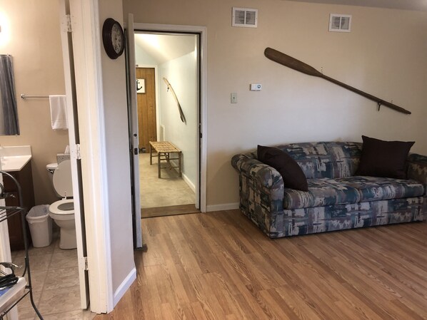 Un salon avec un canapé à motifs, un plancher en bois, une porte donnant sur un couloir et une batte de baseball accrochée au mur.