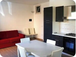 Un espacio habitable pequeño con un sofá rojo, una mesa de comedor con sillas, una zona de cocina con armarios y estufa, y una ventana pequeña.