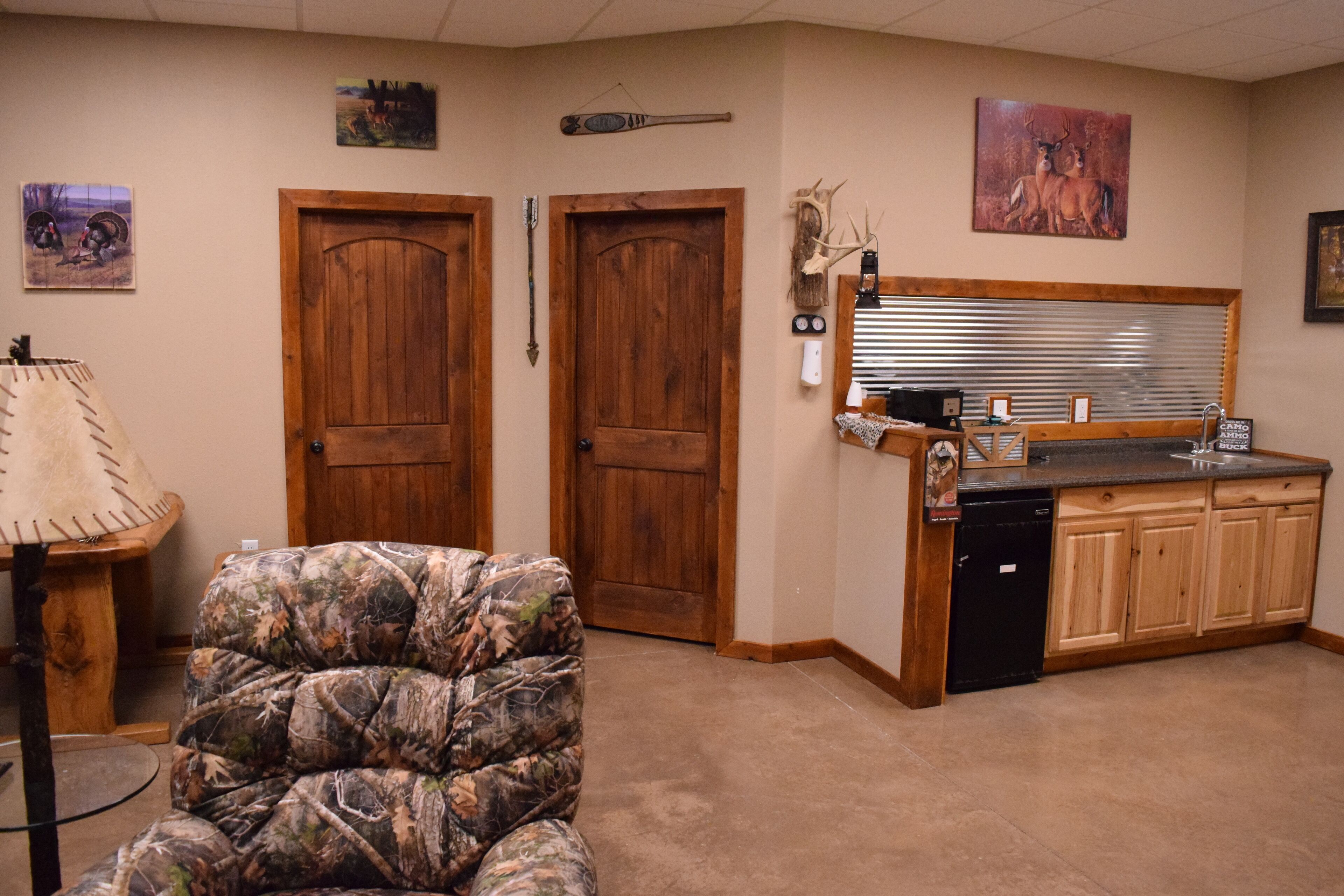 A room with a wooden door, a camouflage chair, a kitchen area with wooden cabinets, and a window with blinds.