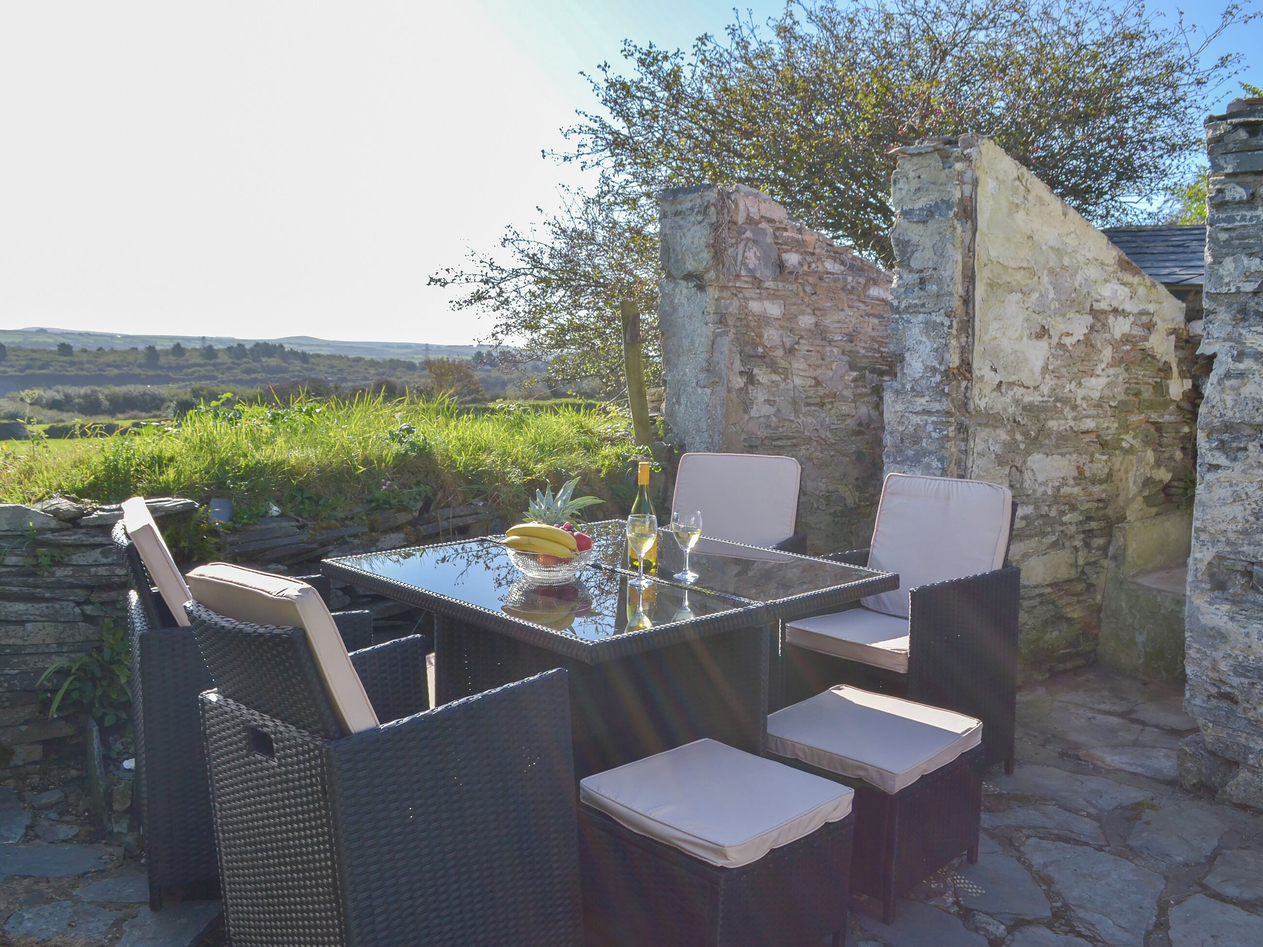 An outdoor dining area with wicker furniture and a glass table, set against a stone wall and lush greenery.