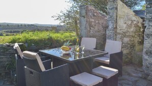 An outdoor dining area with wicker furniture and a glass table, set against a stone wall and lush greenery. - 4 bedroom accommodation in Delabole, near Tintagel (Delabole, near Tintagel)