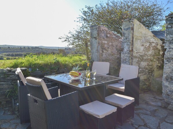 Un área de comedor al aire libre con mobiliario de mimbre y una mesa de vidrio, frente a una pared de piedra y abundante vegetación.