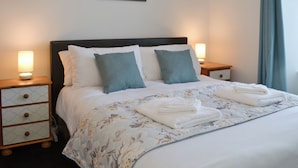 A neatly made bed with a floral comforter, two pillows, and folded towels. There are bedside tables with lamps and a wall-mounted painting above the bed. - 4 bedroom accommodation in Delabole, near Tintagel (Delabole, near Tintagel)
