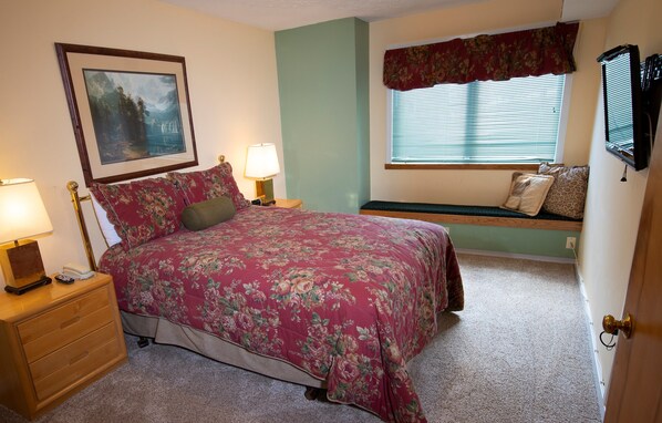 A bedroom with a floral bedspread, a bedside table with a lamp, a nightstand, a window with blinds, and a mounted TV.