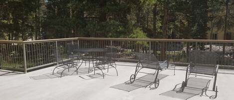 Un patio al aire libre con mesas y sillas de metal, rodeado por una barandilla metálica y árboles.