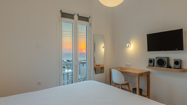 Une chambre d’hôtel équipée d’un lit, d’un bureau, d’une chaise, d’une télévision et d’un balcon offrant une vue sur le coucher du soleil.