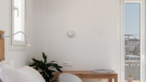 Une chambre à coucher comprenant un lit, un bureau, une chaise, une plante en pot et un balcon avec vue sur les immeubles.