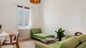 Un salon lumineux avec un canapé vert, une table à manger entourée de chaises et une petite table ronde agrémentée d’une plante.