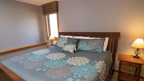 A neatly made bed with patterned bedding, a wooden headboard, two bedside tables with lamps, and a window with blinds. - Charming + Rustic Condo with Ski-in/Ski-Out Access (Ludlow)