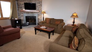 A living room with a stone fireplace, a television mounted above it, and two couches with patterned cushions. - Charming + Rustic Condo with Ski-in/Ski-Out Access (Ludlow)