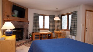 Une chambre à coucher avec un grand lit, un téléviseur fixé au-dessus d’une cheminée, un bureau avec une chaise et une fenêtre avec des rideaux.