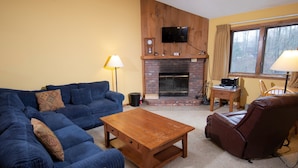 A living room with a blue sofa, a wooden coffee table, a leather armchair, a fireplace, and a television mounted on the wall. - Amazing Location! Charming Ski-in/Ski-out Condo (Ludlow)