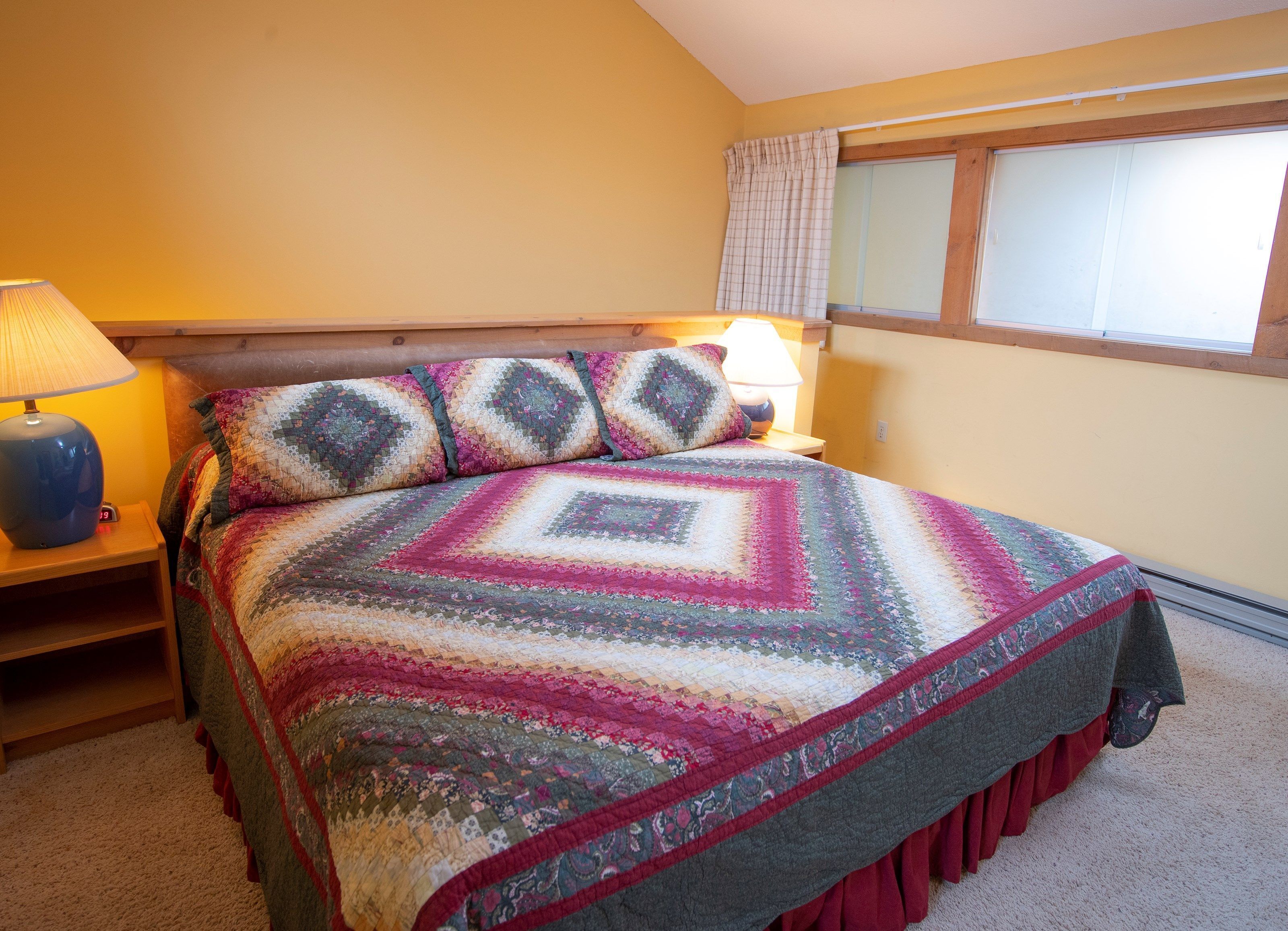 A bedroom with a bed, two bedside lamps, a window with curtains, and a wooden headboard.