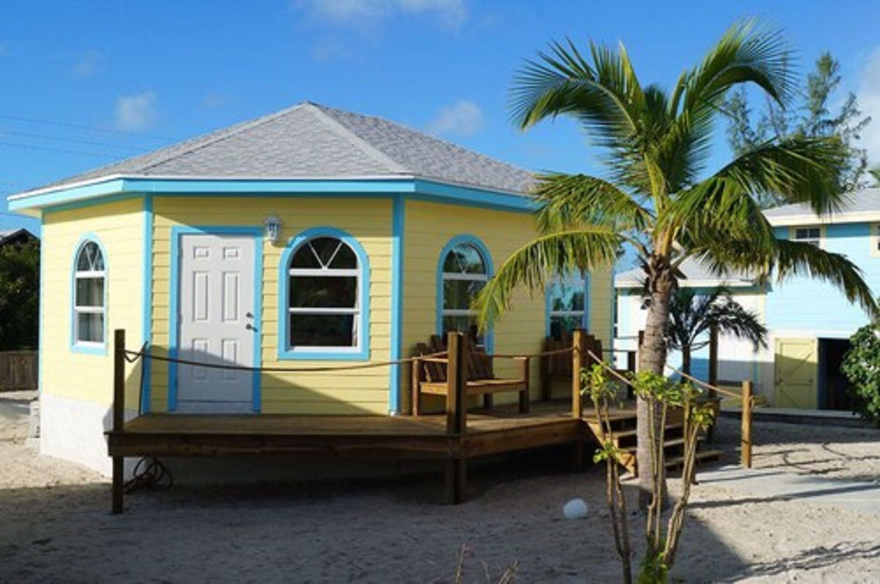 A small, single-story building with a blue and yellow exterior, a wooden deck, and a palm tree in front.