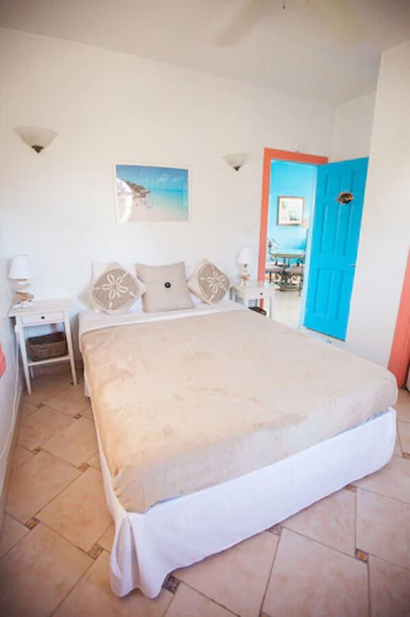 A bedroom with a large bed, a nightstand, a lamp, a painting on the wall, and a blue door.