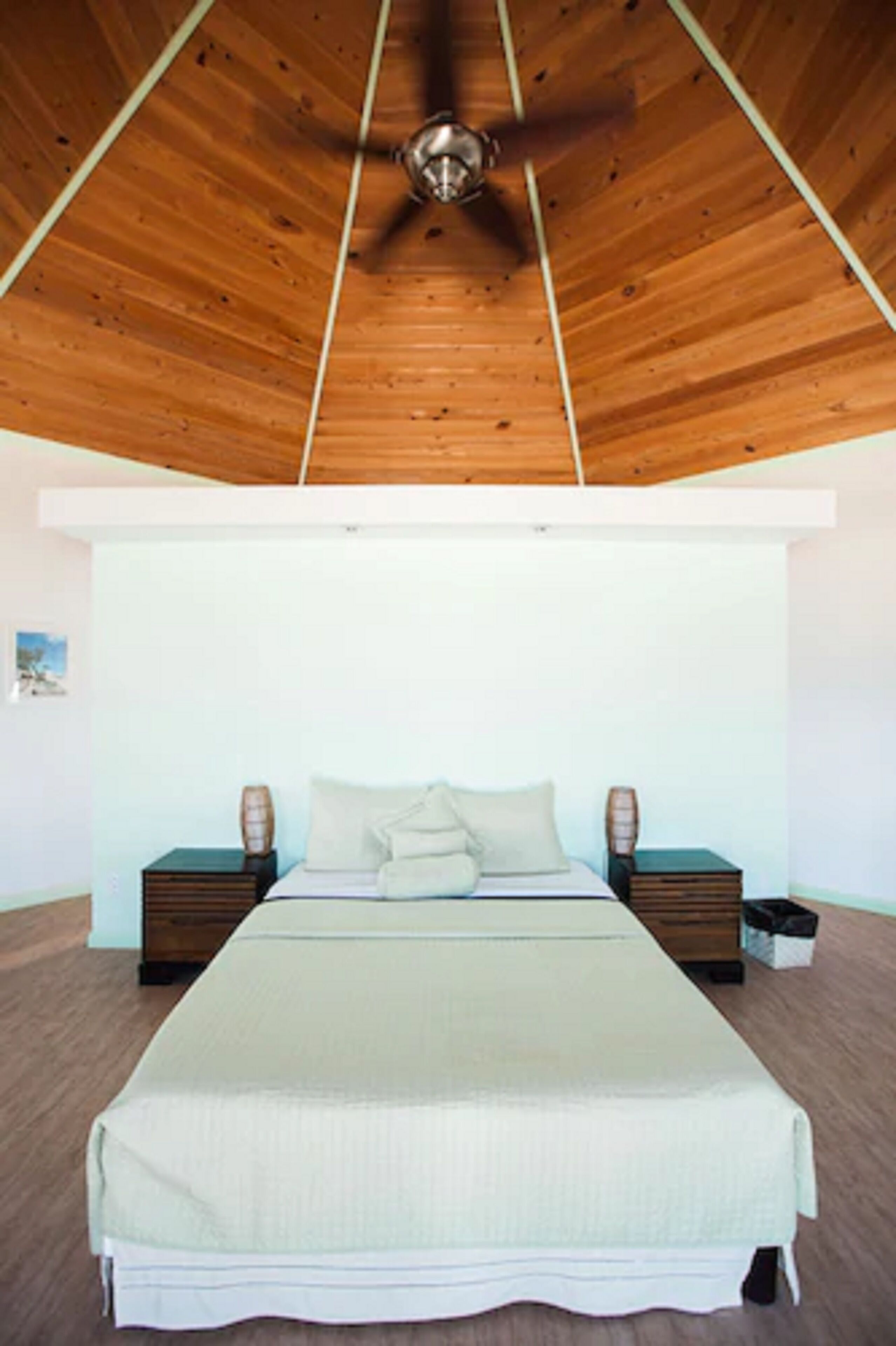 Un dormitorio con techo de madera, una cama con colcha blanca, dos mesitas de noche y un tocador.