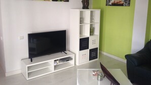 Ein Wohnzimmer mit einem Flachbildfernseher auf einem weißen Standfuß, einer weißen Regaleinheit mit Dekorationselementen, einer grünen Akzentwand und einem schwarzen Ledersofa.