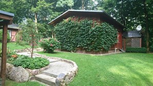 Une maison traditionnelle en bois, dotée d’un toit à pente, entourée d’un jardin bien entretenu avec un chemin de pierre, un petit arbre et des plantes à fleurs.