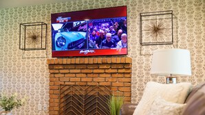 A living room with a brick fireplace, a television mounted above it, patterned wallpaper, and a lamp on a side table. - “Spacious 5-Bedroom Retreat – Perfect for Families & Groups” (Citrus Heights)