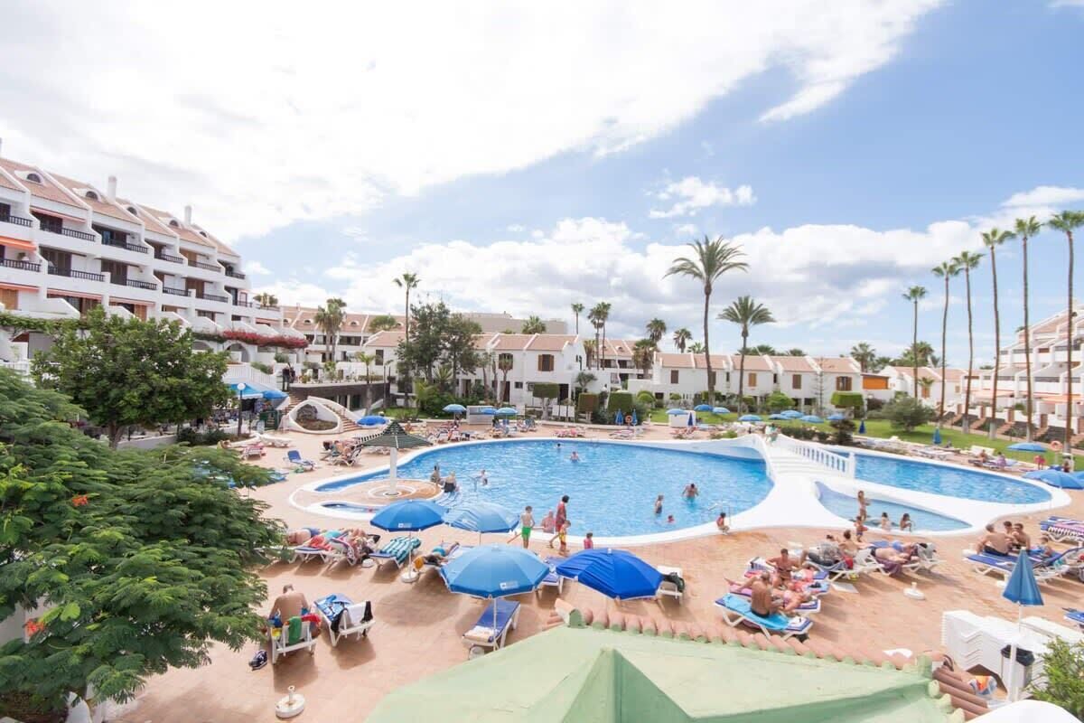 Ein Resort mit einem großen Schwimmbecken, umgeben von Liegestühlen und Sonnenschirmen, Palmen und mehrstöckigen Gebäuden.