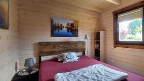A wooden cabin bedroom with a bed, a nightstand, a lamp, a painting, and a window.