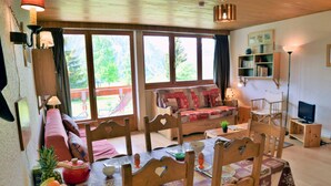 A cozy living room with a wooden ceiling, a sofa, a dining table set with plates and a fruit bowl, and a television on a wooden stand.