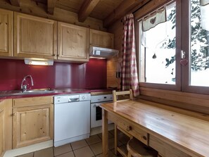 Cocina de cabaña de madera con encimera roja, armarios de madera, lavavajillas blanco y una ventana con cortina de cuadros.