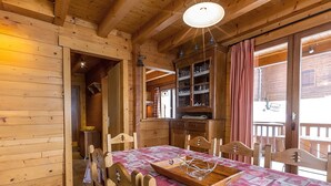 L’intérieur d’un chalet en bois comprend une table à manger, des chaises, un buffet contenant de la verrerie et une fenêtre avec des rideaux.