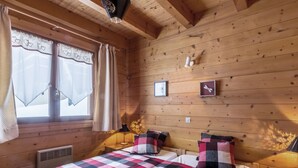 A wooden cabin bedroom with a checkered bedspread, two pillows, a bedside table, a lamp, and a window with curtains. - Cozy chalet for 10, fireplace, garage, WIFI, pets welcome (La Clusaz)